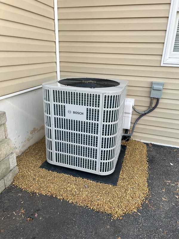 An exterior air conditioning unit with the brand Bosch is installed against a beige siding wall. The unit is placed on a concrete slab and surrounded by gravel.