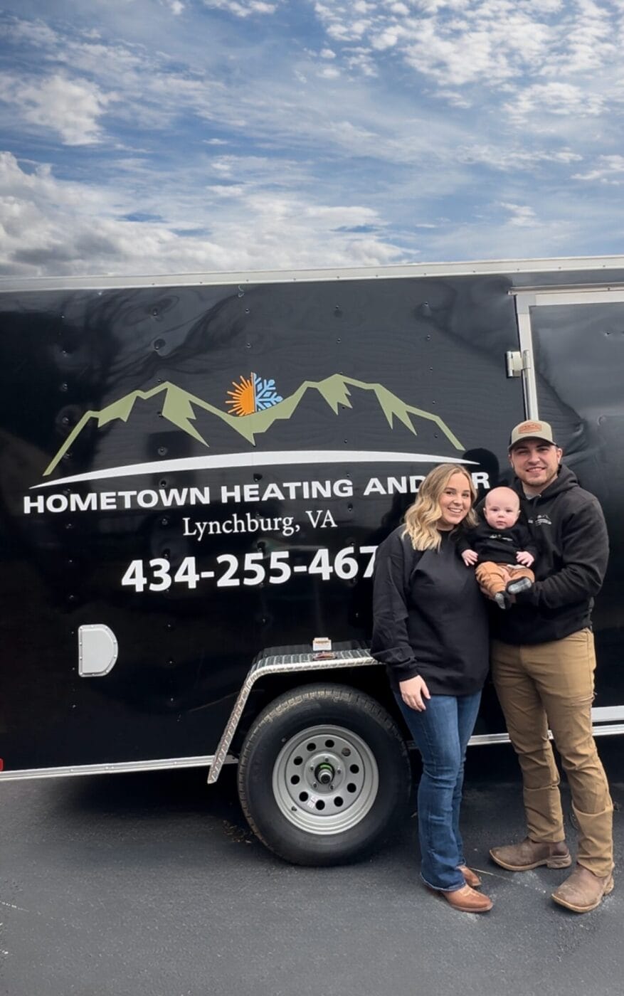 A man and woman, both in black hoodies, stand with a baby in front of a black trailer with Hometown Heating and Air, Lynchburg, VA and a phone number on it, under a partly cloudy sky.