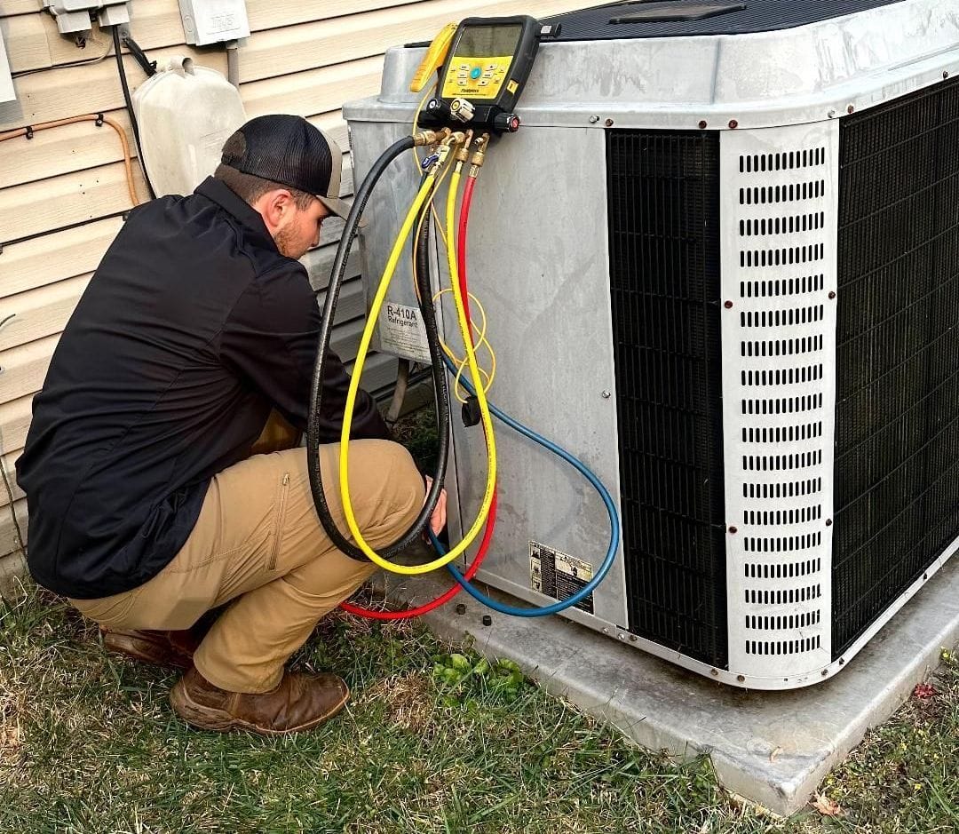 A technician kneels beside an outdoor air conditioning unit, using diagnostic equipment connected with red, blue, and yellow hoses.